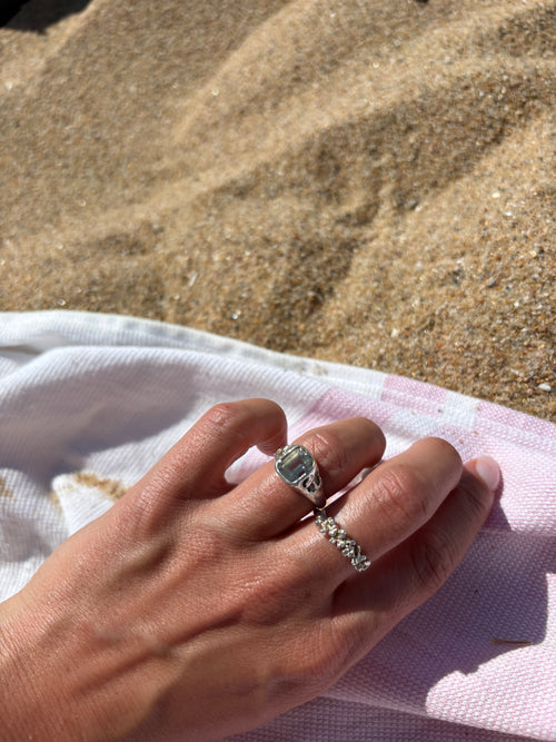 A close-up of a person's hand resting on a pink and white striped towel at the beach, wearing two silver rings, including a textured band ring and a larger signet-style ring.