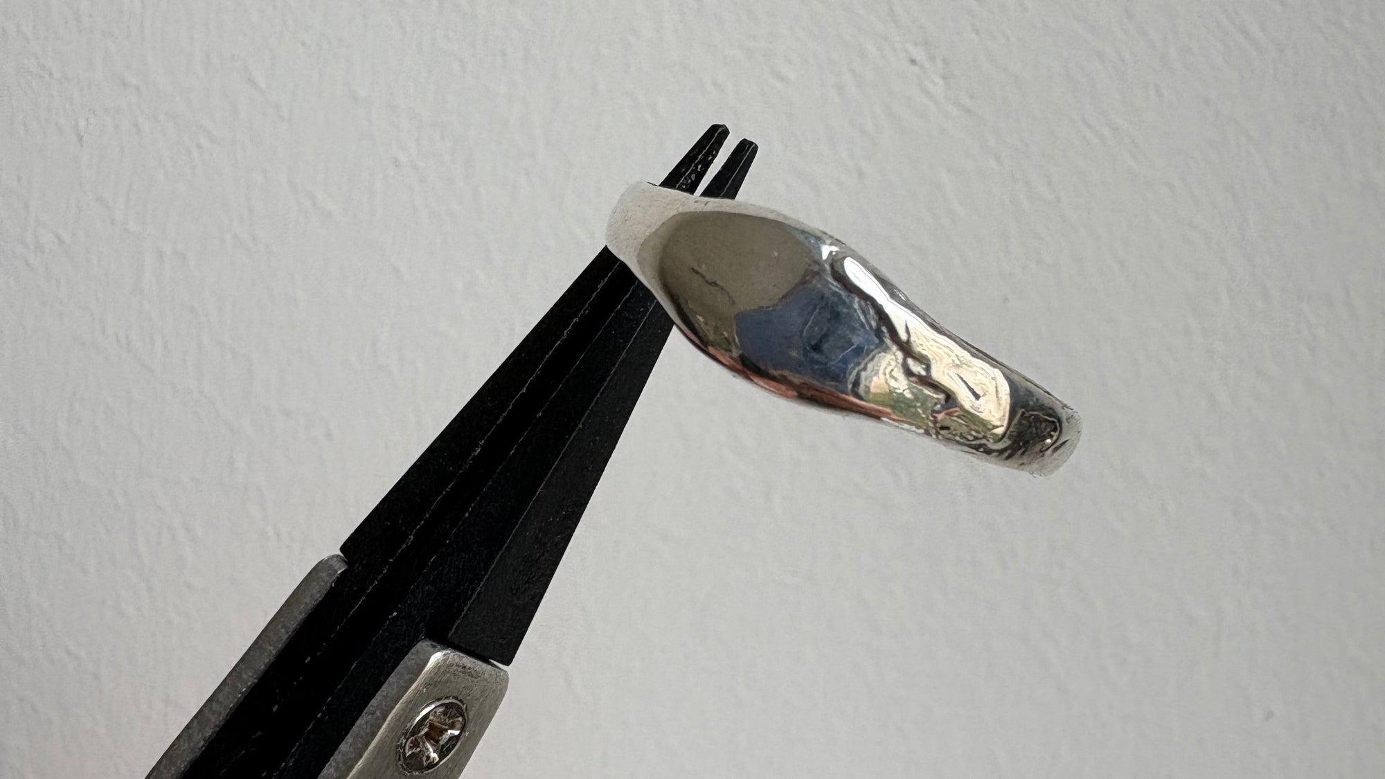A polished customed made silver ring held in a black and silver ring press tool against a plain white textured wall. Made in Amsterdam by Palla Studio.