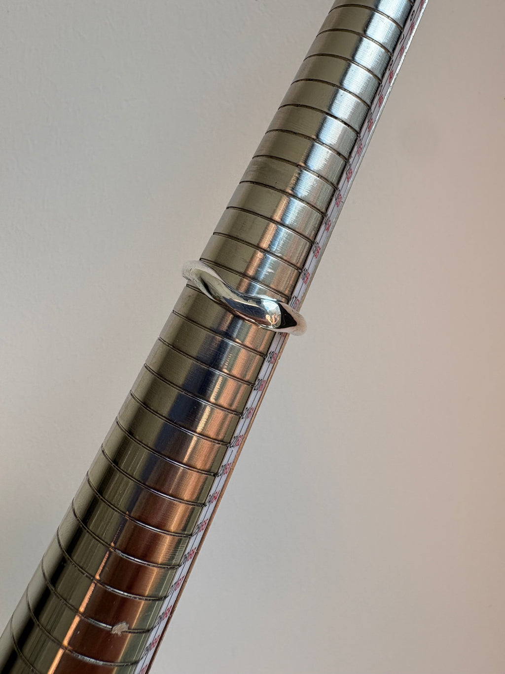 A polished silver metal ring displayed on a metallic measuring mandrel against a plain light gray background.