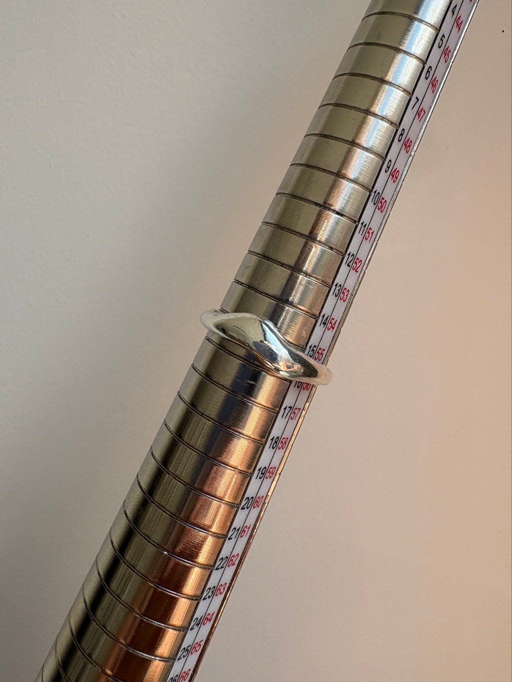 A silver metal ring displayed on a metallic measuring mandrel with printed size markings, against a plain light background.