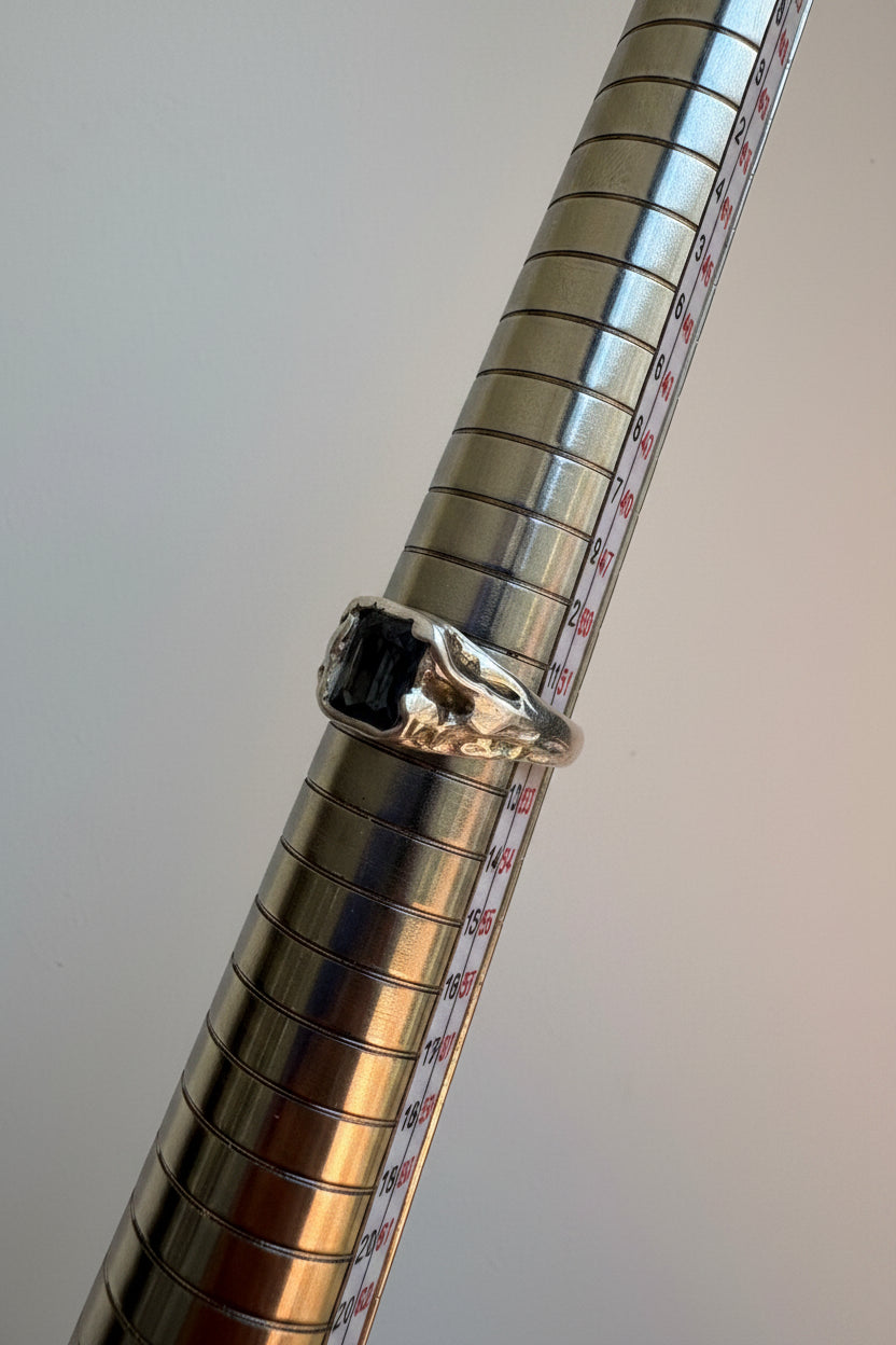 A silver ring with a grey spinel rectangular gemstone displayed on a metallic measuring ring against a plain light background.