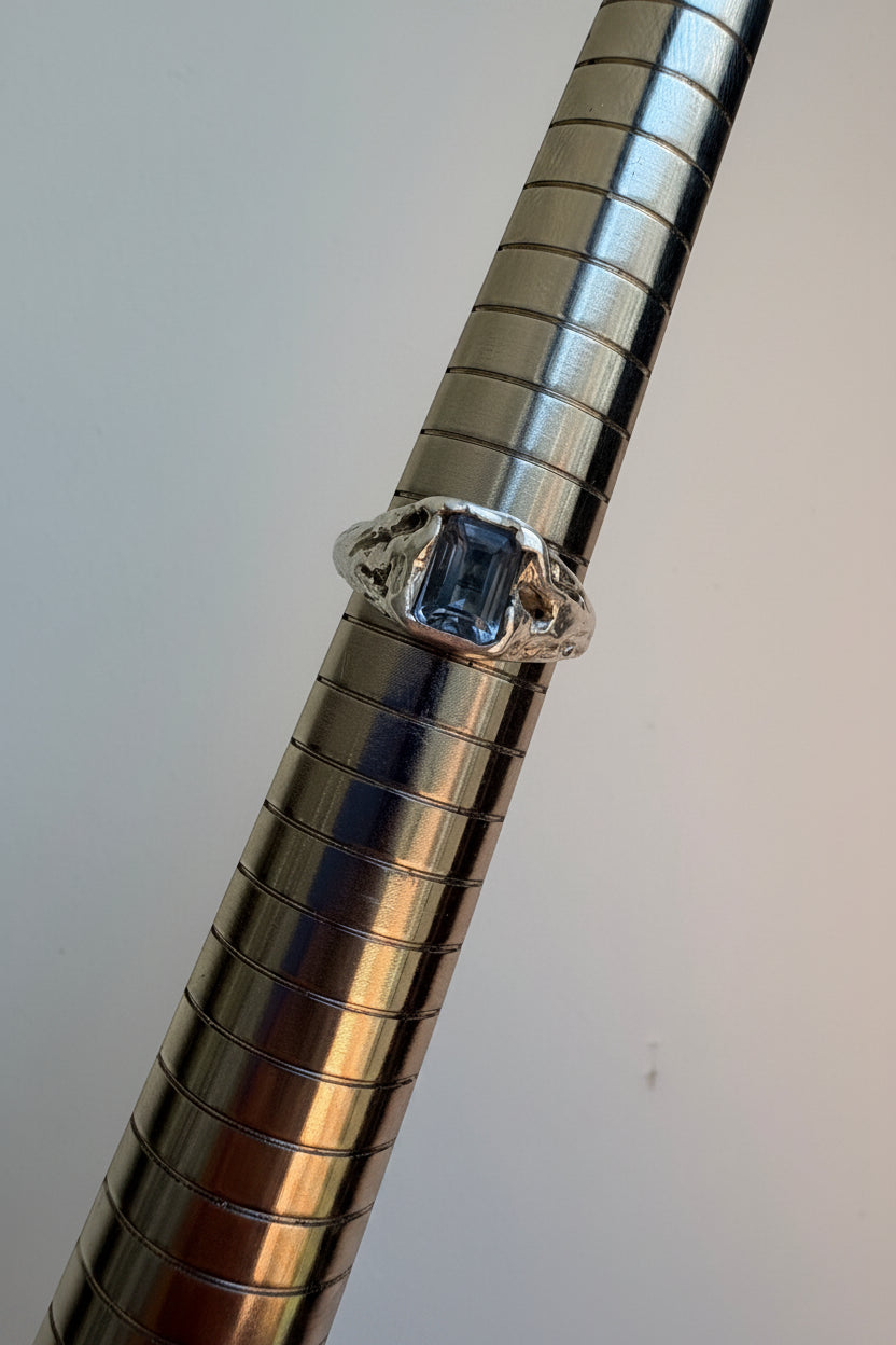 A silver ring with a emerald cut grey spinel, displayed on a metallic ring stand against a plain light background.