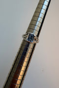 A silver ring with a emerald cut grey spinel, displayed on a metallic ring stand against a plain light background.