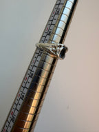 A silver ring with a flowey setting and a rectangular gemstone, shown resting on a metal ring sizer with measurement markings against a plain light background.