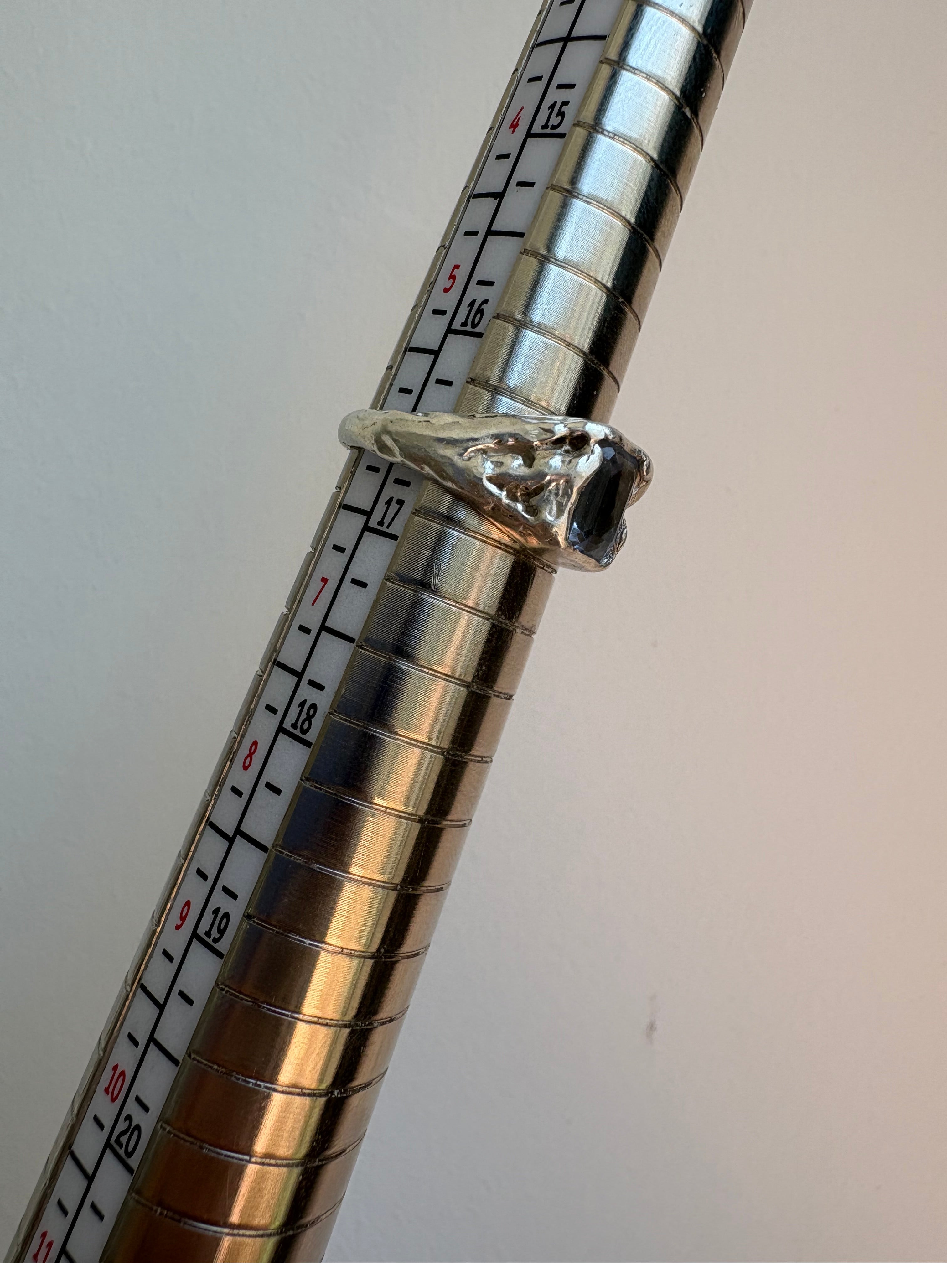 A silver ring with a flowey setting and a rectangular gemstone, shown resting on a metal ring sizer with measurement markings against a plain light background.