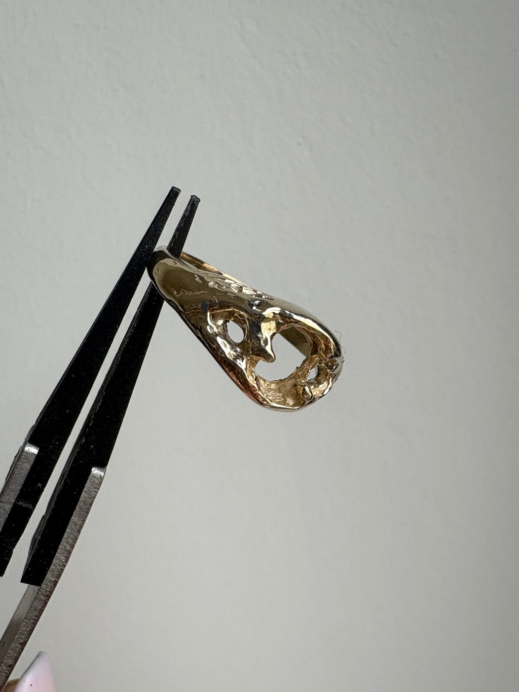 A gold ring with an organic, flowing silhouette is held in forceps against a plain light gray background.