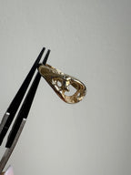 A gold ring with an organic, flowing silhouette is held in forceps against a plain light gray background.