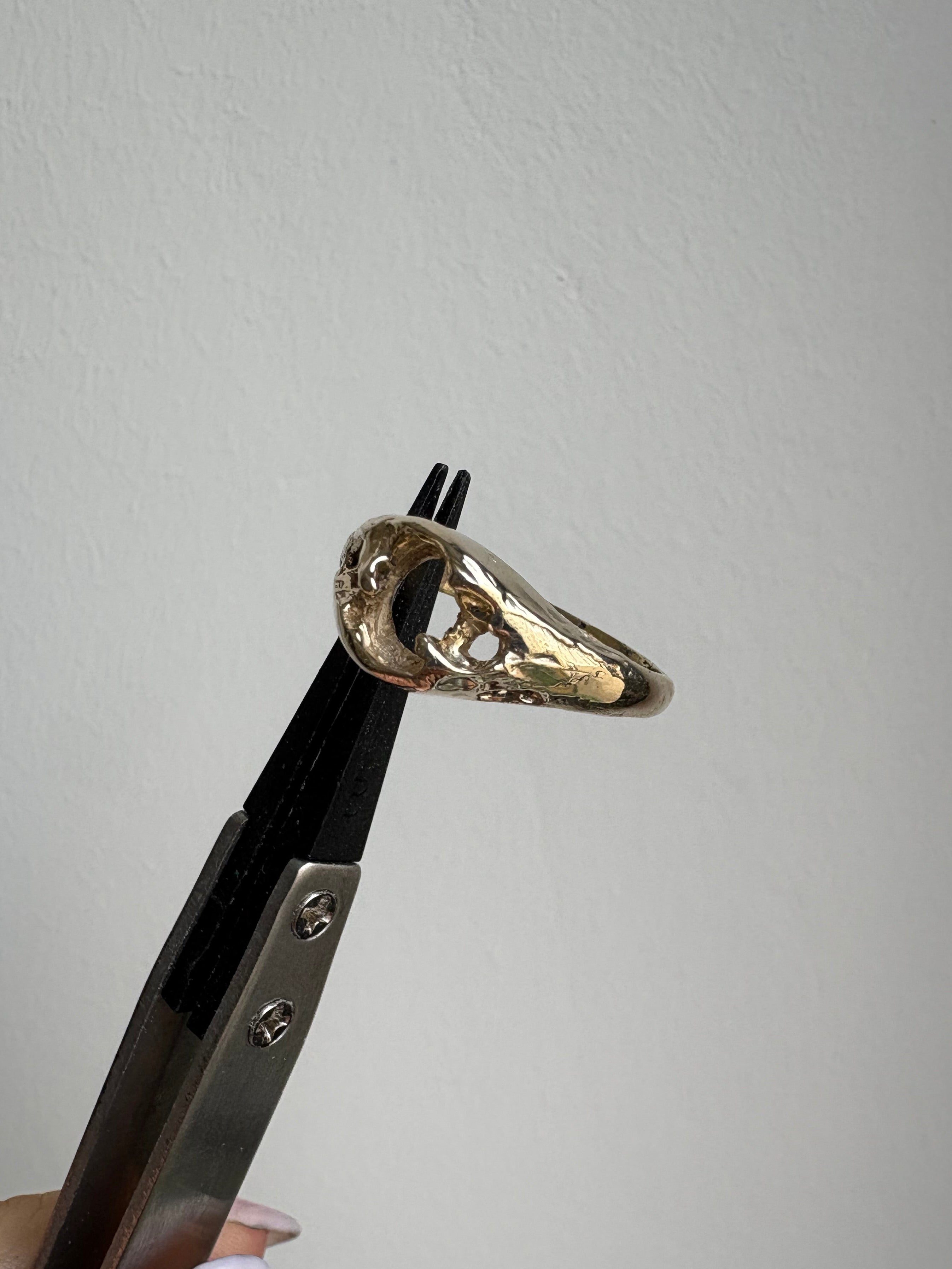 A small metallic object with a gold-toned decorative top and black handle is held in a pair of metal pliers against a plain light gray background. A finger is visible holding the pliers.